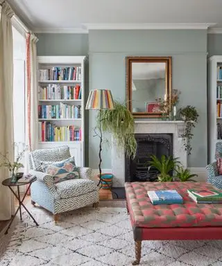 Šedozelený obývací pokoj, bílý krb, zrcadlo na římse, růžový a šedý vzorovaný otoman, knihovna, útulný koberec a křeslo