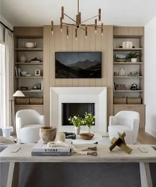 벽난로가 있는 현대적인 거실, 양쪽에 높은 선반, 벽난로 위에 설치된 TV, 크림색 라운지 의자 2개, 플로어 램프, 커피 테이블 및 소파