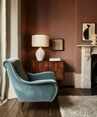 Acogedor salón con paredes pintadas de color rosa apagado, chimenea, aparador de madera oscura, lámpara de mesa color crema y sillón de terciopelo azul