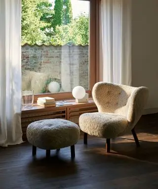 Sillón de piel de oveja y taburete a juego junto a la ventana, aparador bajo decorado con lámpara, libros y adornos, suelo de madera oscura, cortinas transparentes