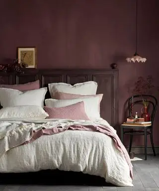 Chambre aux murs peints en rose foncé, regardant un lit double avec une literie blanche, des couvertures roses et des coussins dispersés, une suspension basse au-dessus d'une chaise en bois noire décorée de livres, de fleurs et d'ornements, une tête de lit en bois sombre, un sol gris-brun, une photo encadrée et un feuillage au repos sur la tête de lit.