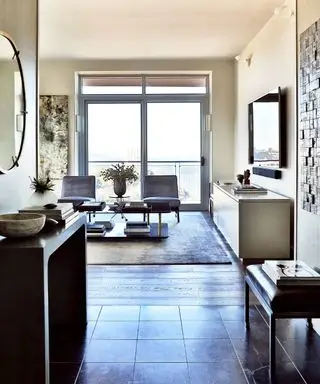 otevřený obývací pokoj, kombinace dlážděných a dřevěných podlah, velký koberec se dvěma křesly, nízký příborník s televizí připevněnou na stěně