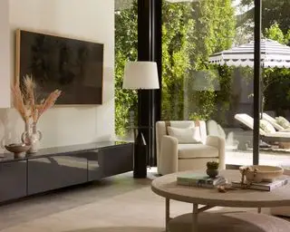 Uvolněný, boho prostor obývacího pokoje, nízký šedý příborník s nástěnnou televizí se světlým dřevěným lemováním umístěným nahoře, krémové křeslo a přírodní koberec, kulatý strukturovaný stupňovitý konferenční stolek, lehátka a slunečník na vnějším pozadí