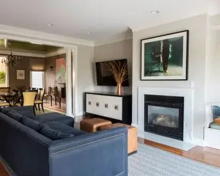 otevřený obývací pokoj a jídelna, dřevěná podlaha s mramorovým krbem, modrá pohovka se dvěma dvojitými koženými podnožkami, sezení ve výklenku, příborník s nástěnnou televizí nahoře, šedý vzorovaný koberec