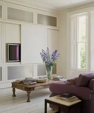 Velký moderní bílý obývací pokoj s bílými umytými palubkami, fialová sedačka, okna s okenicemi, plazmová TV obrazovka skrytá plátnem, dřevěný konferenční stolek