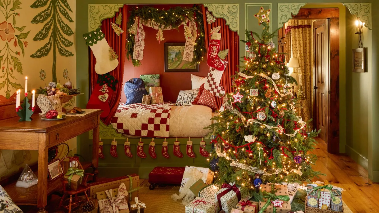 Ein Schlafzimmer mit Bettecke im Six Bells Inn, das mit roter und grüner Folk-Ästhetik gestaltet und weihnachtlich gekleidet ist mit Strümpfen, Patchwork-Accessoires, einem Weihnachtsbaum und verpackten Geschenken' loading='lazy' title=