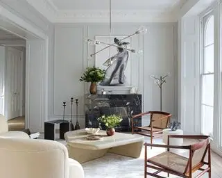 Una sala de estar blanca clásica con sillas de mimbre y madera, iluminación arquitectónica, mesa de centro de piedra y cornisas blancas.