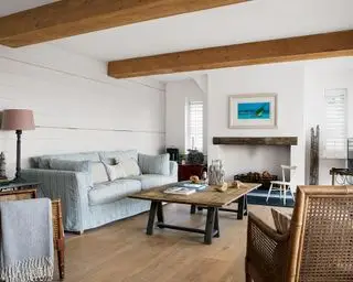 Un salón blanco con vigas de madera, mesa de centro de madera, sillas tejidas y un sofá azul pálido.
