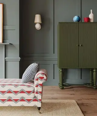 Grønn stue med rødmønstret divan, vegglampe med skjerm på panelmalt vegg, grønt skap