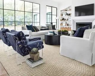 Moderní obývací pokoj s velkým béžovým kobercem, dvěma sladěnými modrými křesly, dvěma bílými pohovkami proti sobě, TV na stěně, police na obou stranách