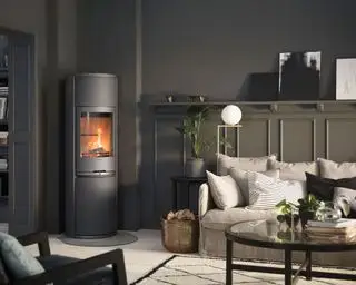 Mørkegråmalet stue med malet vægbeklædning, stor gulvstående contura brændeovn i hjørne af værelset, hvidmalet gulv, hyggelig creme sofa med masser af hynder, gulvlampe ved siden af ​​sofa, mønstret sort og hvidt tæppe, sort og glas sofabord, planter dekoreret rundt i rummet, metal messing kurv til træstammer, sort lænestol i hjørnet af skud