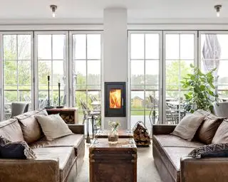 Scandi-inspireret stue med indbygget brændeovn i væggen, plan finish, hyggelig siddeplads med to brune lædersofaer mod hinanden, brune kister, der fungerer som kaffe- og sideborde, logopbevaring i trådet kurv og træskab