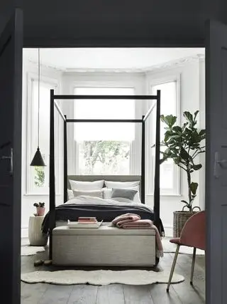 chambre blanche avec lit gris, pouf gris, cadre à baldaquin noir, suspension noire, figuier, fauteuil, accents corail
