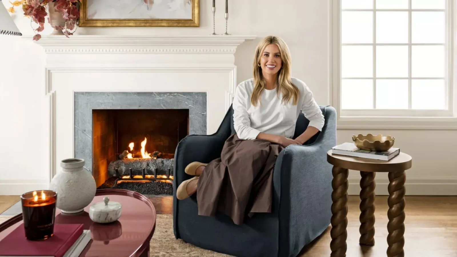 La diseñadora Shea McGee, fotografiada para su colección Target 2025, estaba sentada en un sillón con funda azul marino junto a una chimenea con una mesa auxiliar de madera torneada y una mesa de centro lacada en rojo.' loading='lazy' title=