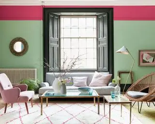 Salon lumineux et coloré avec murs peints en vert et boiseries peintes en rose, grande fenêtre avec volets en bois, parquet en bois clair, tapis crème et rose, canapé gris, fauteuil rose, fauteuil de style osier, table basse en bois et verre, œuvres d'art et miroirs au mur
