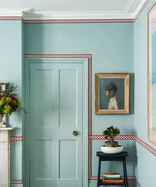 Couloir peint en bleu avec porte peinte en bleu, parquet en bois clair, rayures rouges et planches en damier autour des boiseries. Oeuvre à côté de la porte, tabouret noir avec livres et plantes.