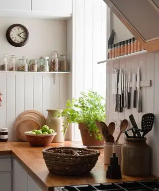 Une cuisine de style rustique, avec des plantes sur le rebord de la fenêtre, des couteaux accrochés au mur et un panier tressé sur le comptoir