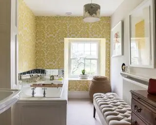 Baño elegante con lavabo y bañera tradicionales, otomana y cómoda antigua, papel pintado amarillo y blanco y gran cesto para la ropa sucia.