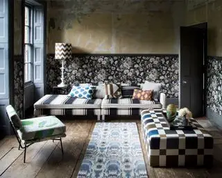 Pared de yeso sin terminar y sala de estar con papel tapiz floral, sillón floral, sofá cama con rayas blancas y negras, otomana cúbica de cuadros metálicos, ventanas de guillotina, pisos desnudos