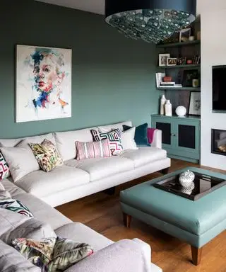 Salón con paredes verdes, suelo de madera y un gran sofá gris en forma de L, otomana de cuero verde que combina con el color de la pintura de las paredes, gran lámpara colgante con bombillas de cristal