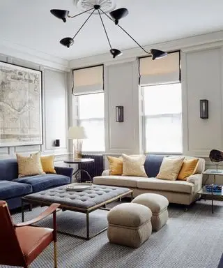 Sala de estar con paredes de color gris pálido, molduras, sofás color crema y azul y alfombra gris, otomana de tela gris oscuro con estructura de metal, dos pufs redondeados grises al lado de la otomana, cojines amarillos, lámpara colgante metálica negra con cinco brazos largos con pantallas.