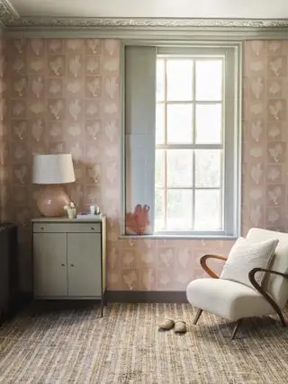 chambre avec papier peint rose, boiseries grises, fauteuil rembourré, petit coffre, lampe, tapis à rayures