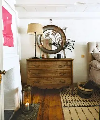 Chambre confortable et champêtre avec murs crème texturés, commode en bois, miroir rond, lampe de table, tapis texturé à motifs, lit dans le coin