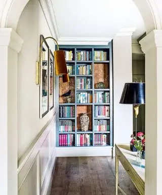 Espace couloir avec murs peints en crème, parquet en bois foncé, console en bois peint en blanc avec lampe de table noire et métallique, donnant sur une bibliothèque intégrée peinte en bleu, remplie de livres et d'ornements, deux tableaux encadrés sur le mur avec des appliques murales à côté