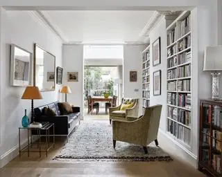 Espace salon et salle à manger décloisonné et lumineux, murs peints en blanc, étagères du sol au plafond remplies de livres, coin salon confortable avec canapé et deux fauteuils, tapis texturé gris doux en dessous, de nombreux tableaux et peintures encadrées sur les murs, lampes de lecture et de bureau à côté coins salons, miroir mural rectangulaire au-dessus du canapé