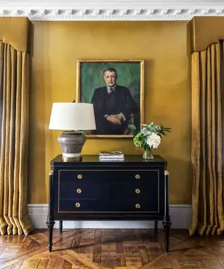 Černá komoda na dřevěné podlaze, stěny zdobené žlutě s obložením a žluté závěsy s garnýžemi