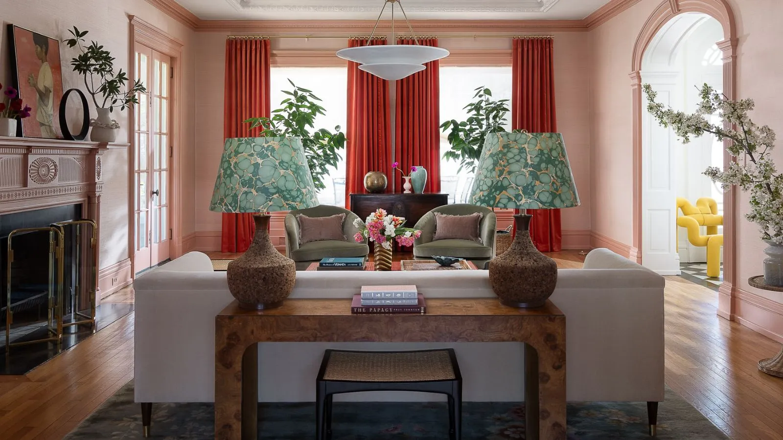 sala de estar rosa abierta con cortinas rojas en la ventana Un sofá blanco con una mesa de consola de madera de burl detrás de él con dos lámparas con tonos de mármol verde' loading='lazy' title=