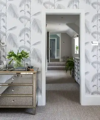 Schlafzimmer mit Teppich, hellgrauer und weißer Tapete mit botanischem Blattmuster und verspiegelter Kommode. Blick entlang eines Flurs im Obergeschoss.