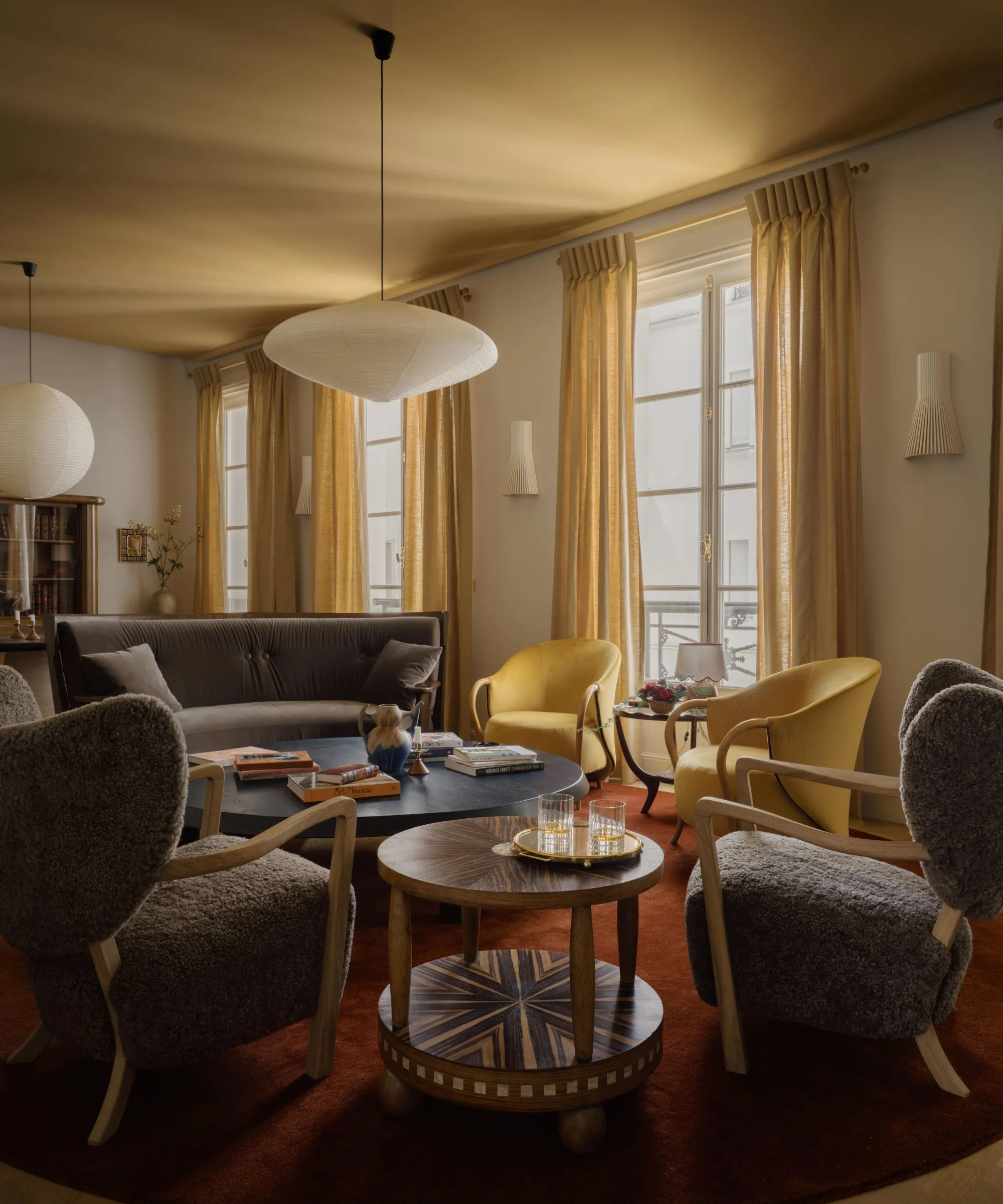 黄土色の天井と温かみのある白い壁、黄土色のカーテンが付いた大きな窓のあるリビング ルーム。ダークレッドのラグとダークニュートラルの椅子。' loading='lazy' title=