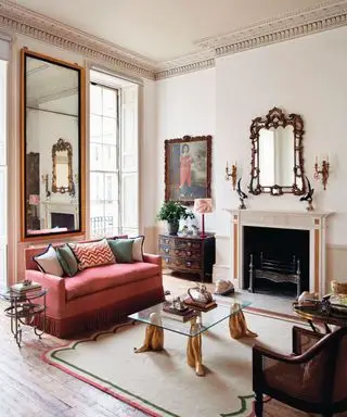 Grand salon avec plafond peint en beige, grand miroir mural, canapé rose, tapis à bords festonnés, cheminée, table basse en verre