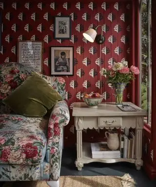 Animada sala de estar con papel pintado rojo, lámpara de pared metálica, gran sillón floral, mesa auxiliar de madera con libros y flores, obras de arte en las paredes