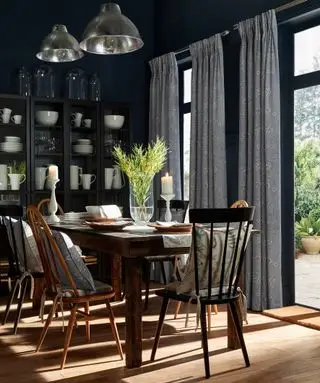 comedor con paredes pintadas de oscuro, piso de madera de tono medio, cortinas estampadas, puertas francesas, vajillas y jarrones color crema