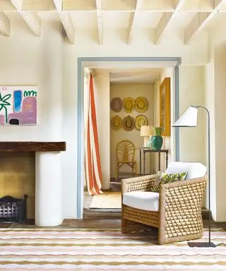 나무 천장 기둥, 고리버들 안락의자, 매끄러운 플로어 램프, 복도를 내다보는 크림색 거실, 줄무늬 커튼, 테이블 램프가 있는 나무 콘솔 테이블, 화려한 러그