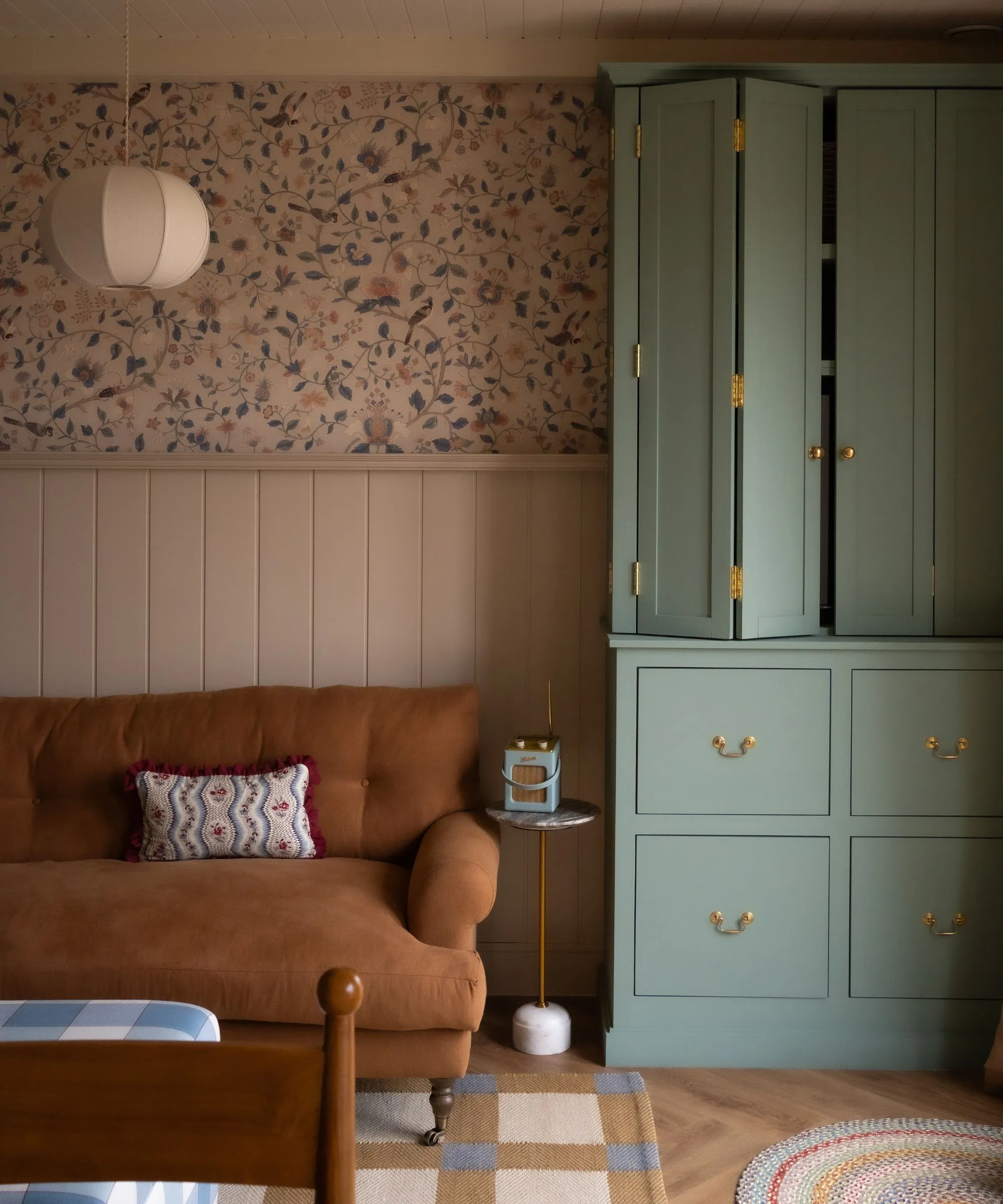 malý útulný priestor s obloženými stenami a polovičnou kvetinovou tapetou, svetlohnedá pohovka a modrá skrinka na mieru, ktorá sa rozkladá a odhaľuje televízor' loading='lazy' title=
