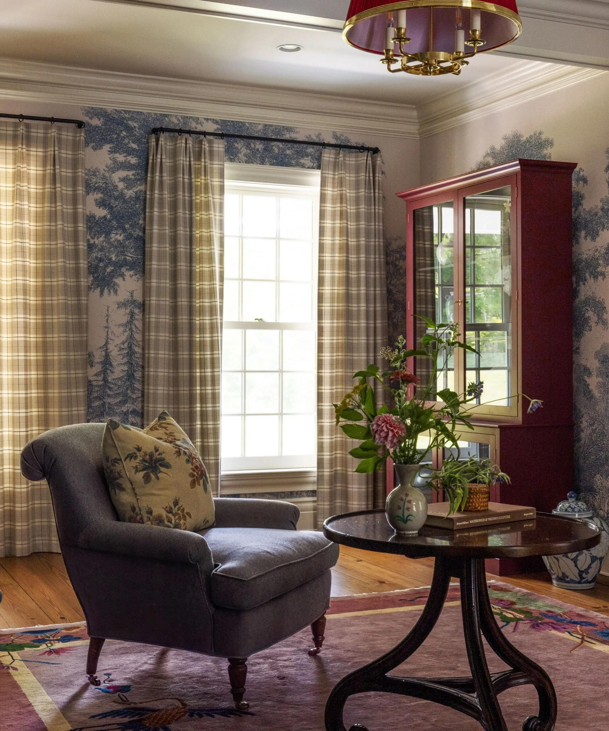 útulná obývačka kreslo a stôl s modro-bielou nástennou maľbou na strom tapeta gingham závesy drevená komoda a kvety vo váze' loading='lazy' title=