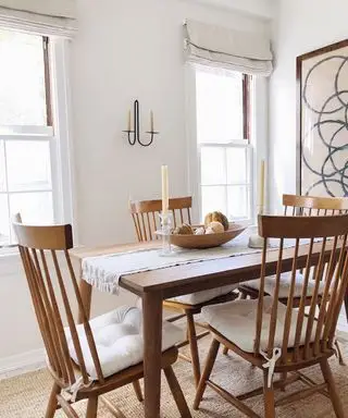 Salle à manger blanche avec table à manger en bois et chaises de salle à manger traditionnelles en bois avec coussins de siège, chemin de table crème avec bougies et bol de fruits en bois, candélabres en métal sur le mur, grande œuvre d'art abstraite