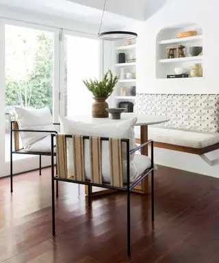 Espace salle à manger décontracté avec murs peints en blanc, parquet en bois foncé, étagères encastrées en arc, décoré d'ornements et d'objets uniques, banc mural avec dossier rembourré, deux chaises de salle à manger palmées avec cadres en métal noir et coussins crème