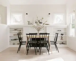 Salle à manger entièrement blanche avec table à manger rectangulaire en bois clair, chaises en bois noir, étagère basse intégrée décorée d'ornements et d'objets à côté de la table, nombreuses fenêtres créant un espace clair et lumineux