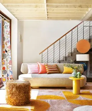 Sala de estar moderna e colorida, clara e brilhante, teto com vigas de madeira clara, sofá creme curvo, tapete estampado amarelo brilhante e laranja escuro, mesa lateral amarela brilhante, almofadas coloridas espalhadas no sofá, cortinas estampadas