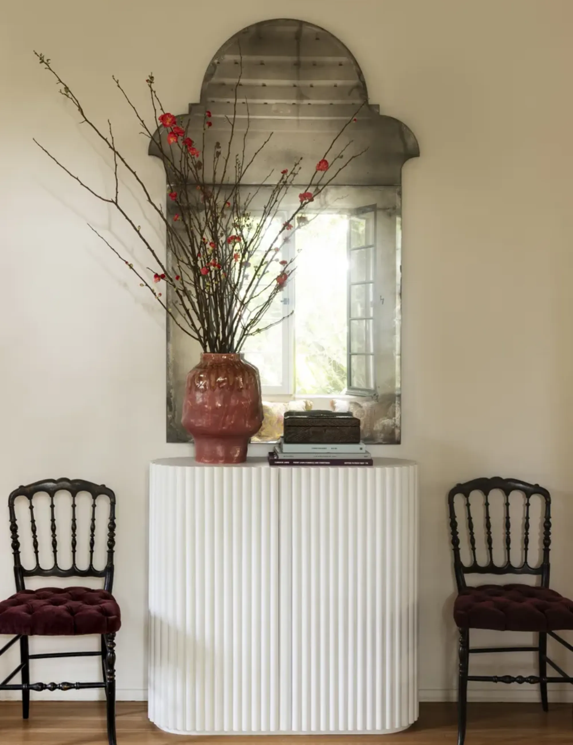 Un miroir antique est suspendu au-dessus d'une console blanche avec deux chaises de salle à manger rembourrées flanquant l'unité.' loading='lazy' title=