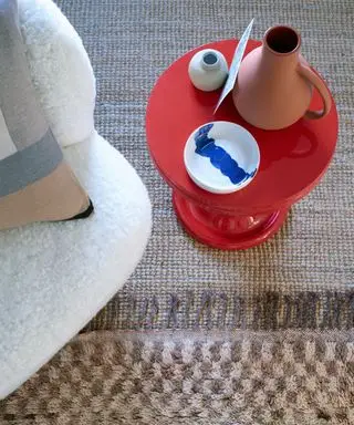 Gros plan d'un tapis à carreaux marron et beige sur un tapis texturé beige, une table d'appoint ronde rouge droite, des ornements sur la table, un coin d'une chaise longue crème avec coussin