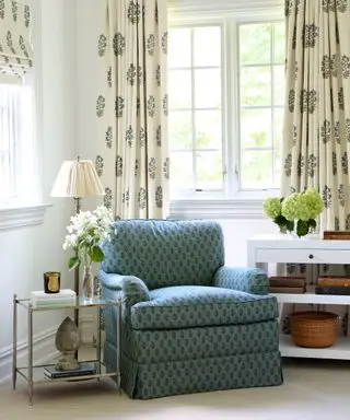 Útulný kout obývacího pokoje s krémovými vzorovanými závěsy, odpovídající roleta, modré křeslo, odkládací stolky s květinami, knihami a ozdobami