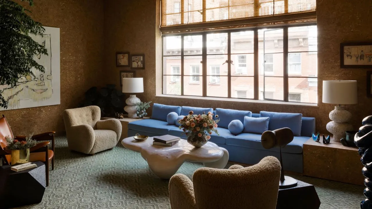 Solbelyst NYC-stue med en overdimensioneret kornblomstblå sofa og khaki-lænestole sammen med et grønt mønstret tæppe og korkbeklædte vægge.' loading='lazy' title=