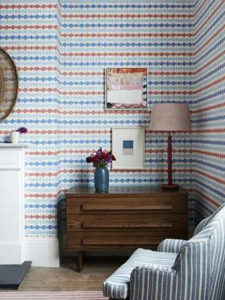 Wohnzimmer mit Geo-Streifentapete in Blau und Rot, gestreifter blauer Sessel, Retro-Kommode, Kunstwerk, Lampe