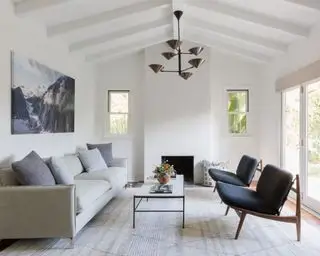 Salon peint en blanc avec lambris et poutres en bois blanc, grand tapis à rayures, canapé crème et deux chaises longues, table basse blanche et noire, suspension métallique, illustration de paysage de montagne