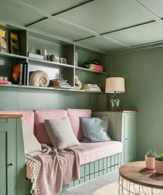 Coin salon avec murs et plafond peints en vert, plafond avec lambris, coin salon en boiseries, fini avec des coussins et des accessoires roses, un tapis arrondi naturel et une table basse, des étagères avec des accessoires décoratifs et des livres